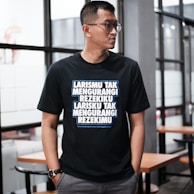 A person wearing glasses stands indoors, dressed in a black T-shirt with bold, white and blue text written in an Indonesian language. The background features glass windows and wooden tables with chairs, suggesting a modern or casual interior setting.