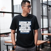 A person wearing glasses stands indoors, dressed in a black T-shirt with bold, white and blue text written in an Indonesian language. The background features glass windows and wooden tables with chairs, suggesting a modern or casual interior setting.