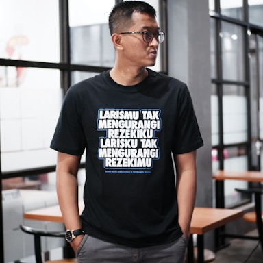 A person wearing glasses stands indoors, dressed in a black T-shirt with bold, white and blue text written in an Indonesian language. The background features glass windows and wooden tables with chairs, suggesting a modern or casual interior setting.