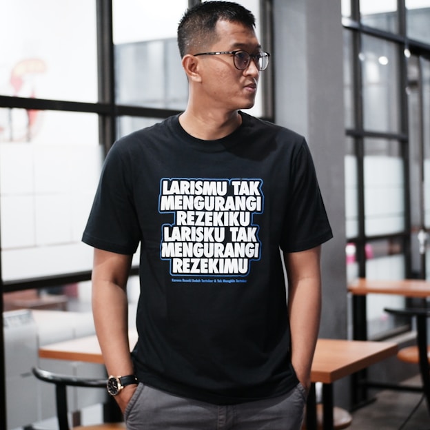 A person wearing glasses stands indoors, dressed in a black T-shirt with bold, white and blue text written in an Indonesian language. The background features glass windows and wooden tables with chairs, suggesting a modern or casual interior setting.