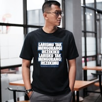 A person wearing glasses stands indoors, dressed in a black T-shirt with bold, white and blue text written in an Indonesian language. The background features glass windows and wooden tables with chairs, suggesting a modern or casual interior setting.
