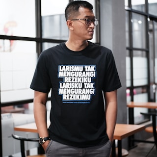 A person wearing glasses stands indoors, dressed in a black T-shirt with bold, white and blue text written in an Indonesian language. The background features glass windows and wooden tables with chairs, suggesting a modern or casual interior setting.