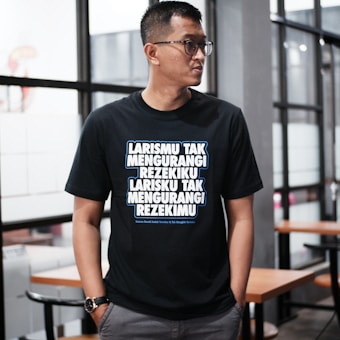 A person wearing glasses stands indoors, dressed in a black T-shirt with bold, white and blue text written in an Indonesian language. The background features glass windows and wooden tables with chairs, suggesting a modern or casual interior setting.