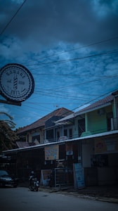 A street view featuring a barbershop sign labeled 'Garage Cuts' with an emblem and the year 2016. The scene includes buildings with tiled roofs and various signage, including a banner for Soto Ayam Lamongan. Power lines crisscross the sky, which is a mix of clouds and blue.