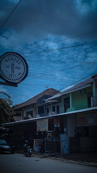 A street view featuring a barbershop sign labeled 'Garage Cuts' with an emblem and the year 2016. The scene includes buildings with tiled roofs and various signage, including a banner for Soto Ayam Lamongan. Power lines crisscross the sky, which is a mix of clouds and blue.
