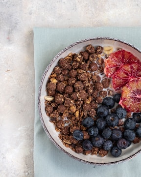 A bowl filled with a mix of cereal clusters, blueberries, and slices of blood orange placed on a textured blue napkin. The bowl is set against a light, neutral-toned background, creating a fresh and inviting composition.