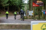 Several cyclists ride along a gravel path surrounded by dense green forest. They wear protective gear, including helmets and jackets. Signs along the trail provide information about the bike trails. The scene conveys a sense of adventure and outdoor activity.
