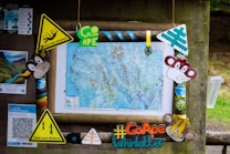 The image features a detailed map mounted on a wooden board, surrounded by various colorful signs and playful decorations. Notable elements include cartoon faces of monkeys, several yellow caution signs, and promotional material labeled #GoApe Whinlatter. The background reveals some greenery, suggesting an outdoor setting.