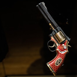 A revolver with intricate design featuring red and gold detailing. The grip is adorned with ornate patterns, and the barrel has a metallic finish. It is displayed against a dark background.