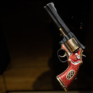 A revolver with intricate design featuring red and gold detailing. The grip is adorned with ornate patterns, and the barrel has a metallic finish. It is displayed against a dark background.