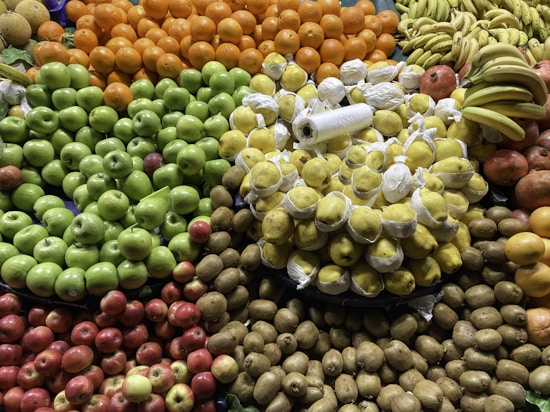 A colorful assortment of various fruits is neatly arranged in a market display. Bright oranges, vibrant green apples, lemons wrapped in protective sheets, bananas, and kiwi are prominently featured.