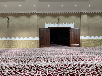 A well-lit, ornately designed interior with a patterned red carpet covering the floor. The beige-colored walls have decorative white trim and stone accents at the lower portion. In the center, there are large wooden doors, currently open, revealing a darkened room inside. Small light fixtures are mounted on the walls, and recessed ceiling lights illuminate the space.