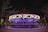 A carousel lit up with vibrant purple and white lights in a nighttime setting. The carousel features various intricately carved animals. Bare tree branches are visible in the background, suggesting a winter or autumn season.