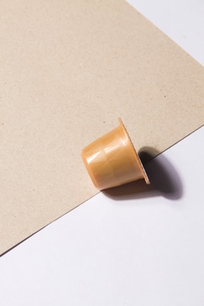 A beige coffee capsule rests on a surface, partially on a light brown textured paper and white background. The capsule casts a soft shadow due to the lighting.