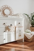 A cozy and tidy nursery corner features a white chest of drawers with three handles. On top of it lies a rolled-up baby blanket with a subtle pattern and a decorative circular mirror with a woven frame. A small plush toy and a cloth book hang from a knob. A wicker basket filled with more baby blankets sits beside the drawers, with a potted plant adding greenery to the scene. Sunlight streams through, casting soft shadows on the wooden floor.