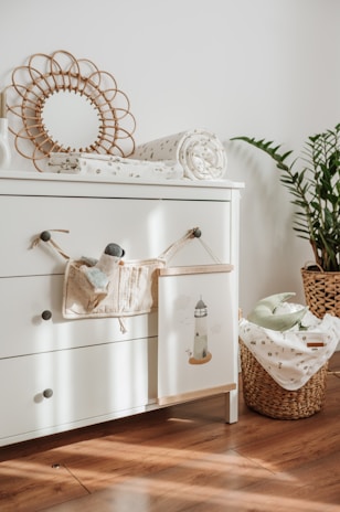 A cozy and tidy nursery corner features a white chest of drawers with three handles. On top of it lies a rolled-up baby blanket with a subtle pattern and a decorative circular mirror with a woven frame. A small plush toy and a cloth book hang from a knob. A wicker basket filled with more baby blankets sits beside the drawers, with a potted plant adding greenery to the scene. Sunlight streams through, casting soft shadows on the wooden floor.