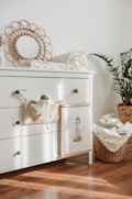 A cozy and tidy nursery corner features a white chest of drawers with three handles. On top of it lies a rolled-up baby blanket with a subtle pattern and a decorative circular mirror with a woven frame. A small plush toy and a cloth book hang from a knob. A wicker basket filled with more baby blankets sits beside the drawers, with a potted plant adding greenery to the scene. Sunlight streams through, casting soft shadows on the wooden floor.