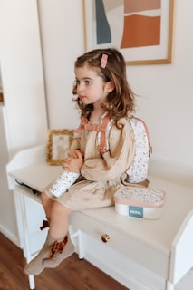 A young child with light brown curly hair, sitting on a white desk. The child is wearing a beige dress, a pink hair clip, and a floral-patterned backpack. She is holding a matching water bottle and a small box with the label 'MEPAL'. The scene is cozy, with a framed abstract art piece on the wall and a wooden floor.
