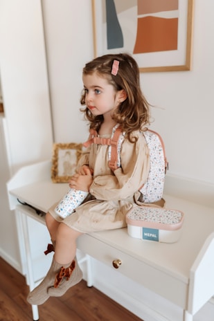 A young child with light brown curly hair, sitting on a white desk. The child is wearing a beige dress, a pink hair clip, and a floral-patterned backpack. She is holding a matching water bottle and a small box with the label 'MEPAL'. The scene is cozy, with a framed abstract art piece on the wall and a wooden floor.