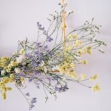 A delicate arrangement of small, multi-colored flowers is hanging on a wall. The bouquet includes light yellow, white, and purple flowers held together in a decorative triangular wooden frame.
