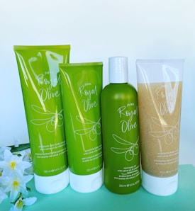 Four beauty product containers are displayed against a plain background. The containers are green and white, labeled 'JAFRA Royal Olive,' with illustrations of olives on them. There are flowers placed beside the containers for decoration.