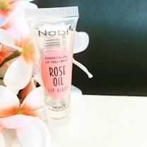 A tube of Nabi Essential Oil Lip Treatment labeled as Rose Oil Lip Gloss stands upright against a soft black background. The tube is transparent with pink labeling and floral imagery on the left side adds an elegant touch to the composition.