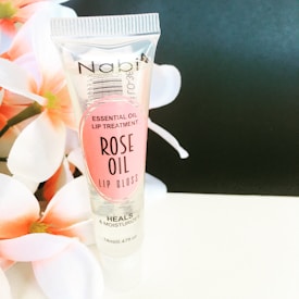 A tube of Nabi Essential Oil Lip Treatment labeled as Rose Oil Lip Gloss stands upright against a soft black background. The tube is transparent with pink labeling and floral imagery on the left side adds an elegant touch to the composition.