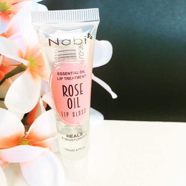 A tube of Nabi Essential Oil Lip Treatment labeled as Rose Oil Lip Gloss stands upright against a soft black background. The tube is transparent with pink labeling and floral imagery on the left side adds an elegant touch to the composition.