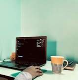 A workspace featuring an open laptop on a white desk, with a person’s hand using a mouse alongside. A distinctive orange and black checkered cup and a closed notebook with a pen rest nearby. The background wall is painted in a calming aqua color.