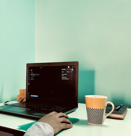 A workspace featuring an open laptop on a white desk, with a person’s hand using a mouse alongside. A distinctive orange and black checkered cup and a closed notebook with a pen rest nearby. The background wall is painted in a calming aqua color.