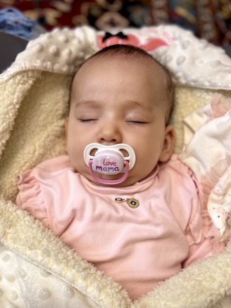 A baby is sleeping peacefully wrapped in a soft, textured blanket. The baby is wearing a pink outfit and has a pacifier in its mouth reading 'I love mama.' The background is slightly blurred with some colorful patterns.