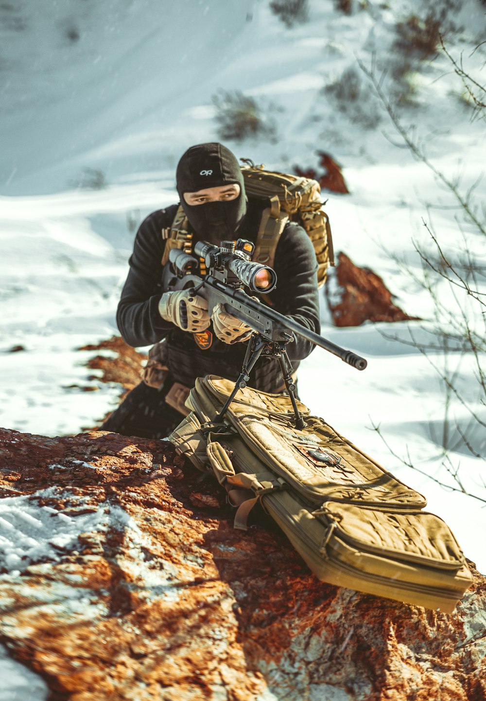 a man with a rifle sitting on a rock in the snow