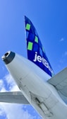 A close-up view of an airplane's tail fin with the JetBlue logo prominently displayed against a clear blue sky. The tail features a geometric design in blue and green squares. The airplane's rear fuselage is visible, capturing details of its sleek, metallic surface.