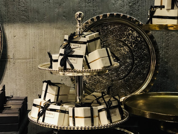 A decorative two-tiered stand holds multiple small, elegant white boxes with black ribbons and gold accents, labeled 'Le Rêve'. The stand is ornate with a metallic finish and is set against a textured, dark background with intricate metal trays to the side, conveying a luxurious and sophisticated atmosphere.