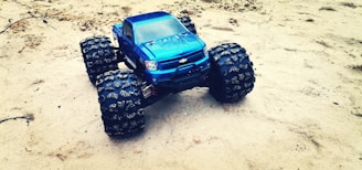A blue toy truck with large, rugged tires is placed on a sandy surface. The vehicle is detailed with a visible front grille and headlights, and the tires are covered with sand, indicating movement in rough terrain.