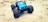 A blue toy truck with large, rugged tires is placed on a sandy surface. The vehicle is detailed with a visible front grille and headlights, and the tires are covered with sand, indicating movement in rough terrain.