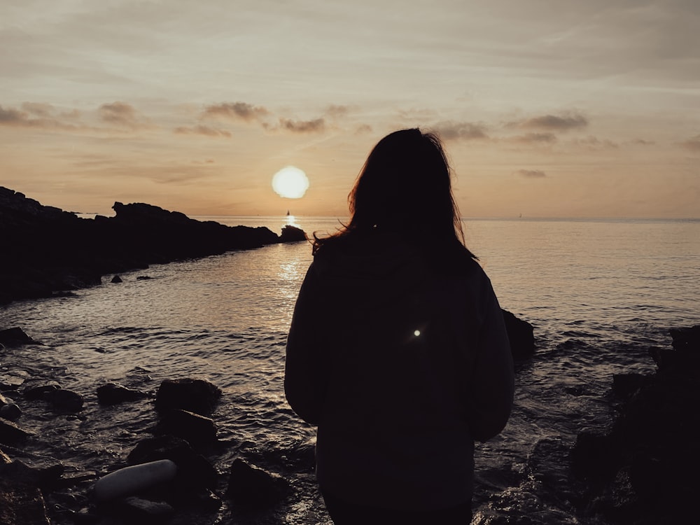 Eine Frau steht an einem felsigen Strand und beobachtet den Sonnenuntergang