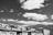 A black and white photograph featuring a series of apartment buildings with flat roofs and multiple windows. The sky is filled with numerous fluffy clouds, and there are hills in the background covered in dense foliage. The buildings have a distinct rectangular shape, and there is some visible laundry hanging outside on one of the balconies.