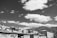 A black and white photograph featuring a series of apartment buildings with flat roofs and multiple windows. The sky is filled with numerous fluffy clouds, and there are hills in the background covered in dense foliage. The buildings have a distinct rectangular shape, and there is some visible laundry hanging outside on one of the balconies.