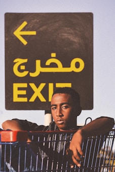 A young person leans on a shopping cart with a serious expression against the backdrop of an exit sign. The sign displays the word 'EXIT' in both Arabic and English, with a large yellow arrow pointing left.