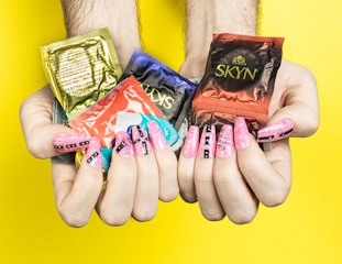 Hands with long, painted nails holding several colorful condom packages against a bright yellow background. The nails are decorated with pink and black designs.