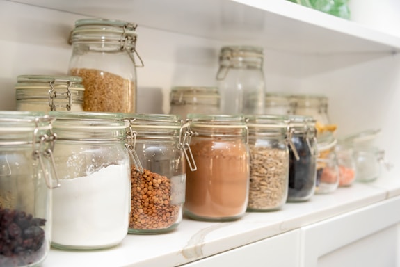 Glass jars filled with various dry ingredients are neatly arranged on white shelves. The jars, which have metal clasp lids, contain items such as flour, grains, seeds, and dried fruits. The arrangement is orderly, creating a sense of organization and tidiness within a kitchen or pantry setting.