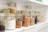 Glass jars filled with various dry ingredients are neatly arranged on white shelves. The jars, which have metal clasp lids, contain items such as flour, grains, seeds, and dried fruits. The arrangement is orderly, creating a sense of organization and tidiness within a kitchen or pantry setting.