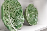 Two ceramic plates designed to resemble large green cabbage leaves are displayed on a white shelf. The plates have intricate vein patterns and are mounted on stands.