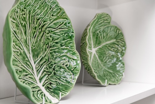 Two ceramic plates designed to resemble large green cabbage leaves are displayed on a white shelf. The plates have intricate vein patterns and are mounted on stands.