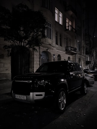 A dark street scene at night features a parked black SUV in front of an old building with several windows lit up from inside. The street is lined with trees, and the atmosphere is quiet and still.