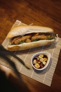 A sandwich filled with fried ingredients and vegetables is placed on a bamboo mat. Next to the sandwich is a small white dish containing a dark sauce with slices of garlic and chili.