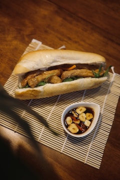 A sandwich filled with fried ingredients and vegetables is placed on a bamboo mat. Next to the sandwich is a small white dish containing a dark sauce with slices of garlic and chili.