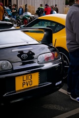 A collection of cars is parked in an outdoor setting. A prominent black Toyota Supra with a yellow license plate is in the foreground. Another brightly colored car, possibly a yellow sports car, is visible alongside. Several people are gathered around the cars, with some admiring or engaged in conversation. The scene conveys an automotive gathering or car show atmosphere.