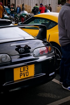 A collection of cars is parked in an outdoor setting. A prominent black Toyota Supra with a yellow license plate is in the foreground. Another brightly colored car, possibly a yellow sports car, is visible alongside. Several people are gathered around the cars, with some admiring or engaged in conversation. The scene conveys an automotive gathering or car show atmosphere.